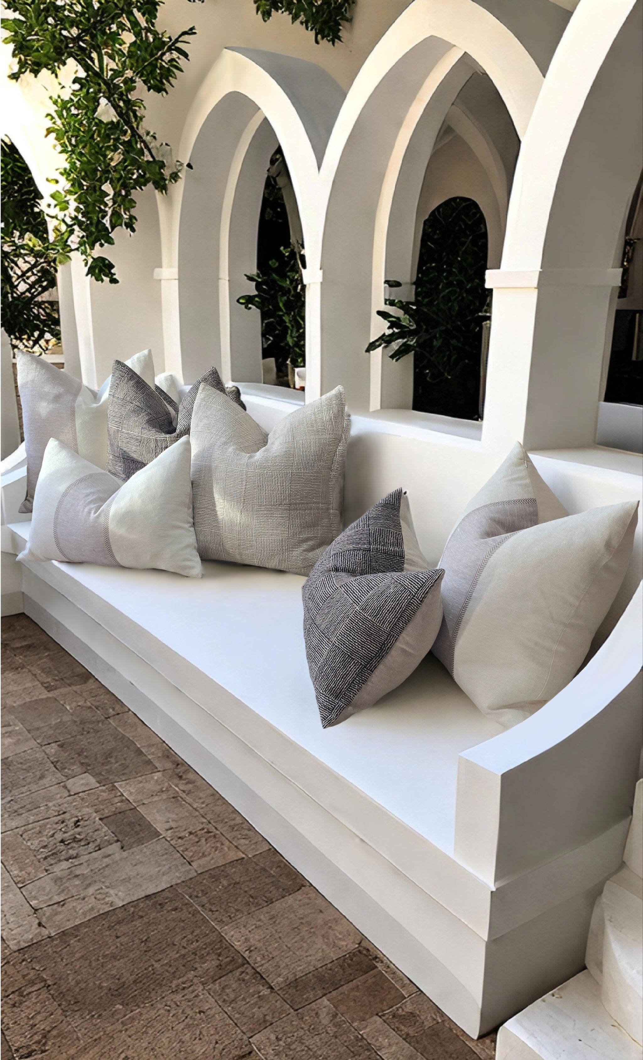 White bench with gray and white pillows in an outdoor setting with arched stone walls.