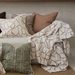 Living room with a beige sofa adorned with patterned cushions and a blanket, against a neutral wall with a shelf.