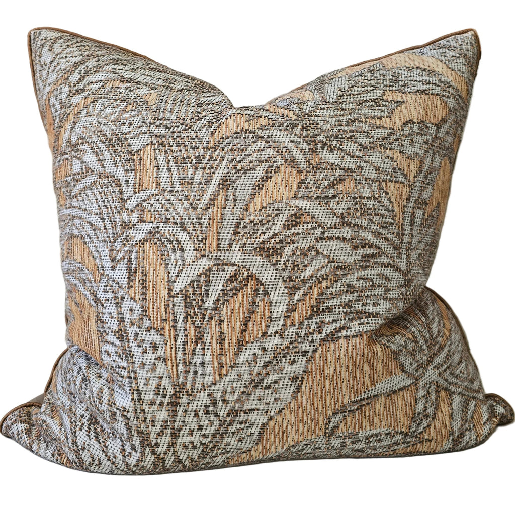 A square decorative throw pillow with a woven botanical pattern in beige, brown, and gray, resting on a light beige sofa. In the background, a wooden shelf holds decorative vases and textured spherical ornaments against a warm terracotta wall.
