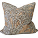 A square decorative throw pillow with a woven botanical pattern in beige, brown, and gray, resting on a light beige sofa. In the background, a wooden shelf holds decorative vases and textured spherical ornaments against a warm terracotta wall.